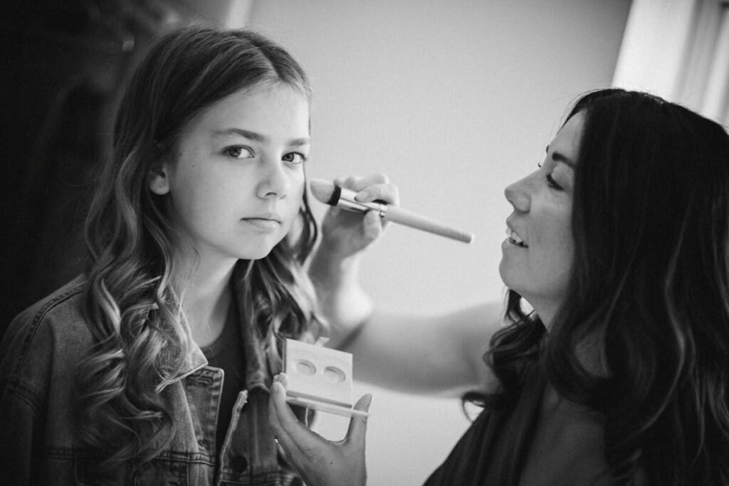 A girl receives a makeup touch-up from an artist in a black and white portrait.