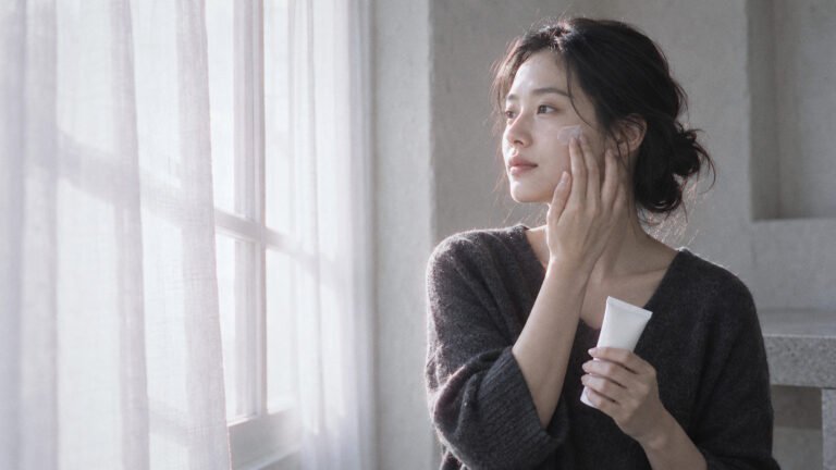 Young woman by a bright window smoothing sunscreen onto her cheekbone in soft morning light