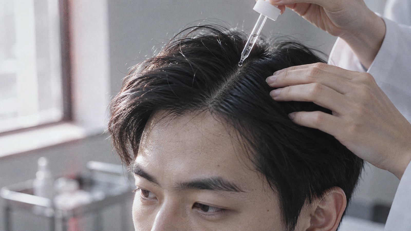 Glass dropper applying a clear serum along a man's hair parting