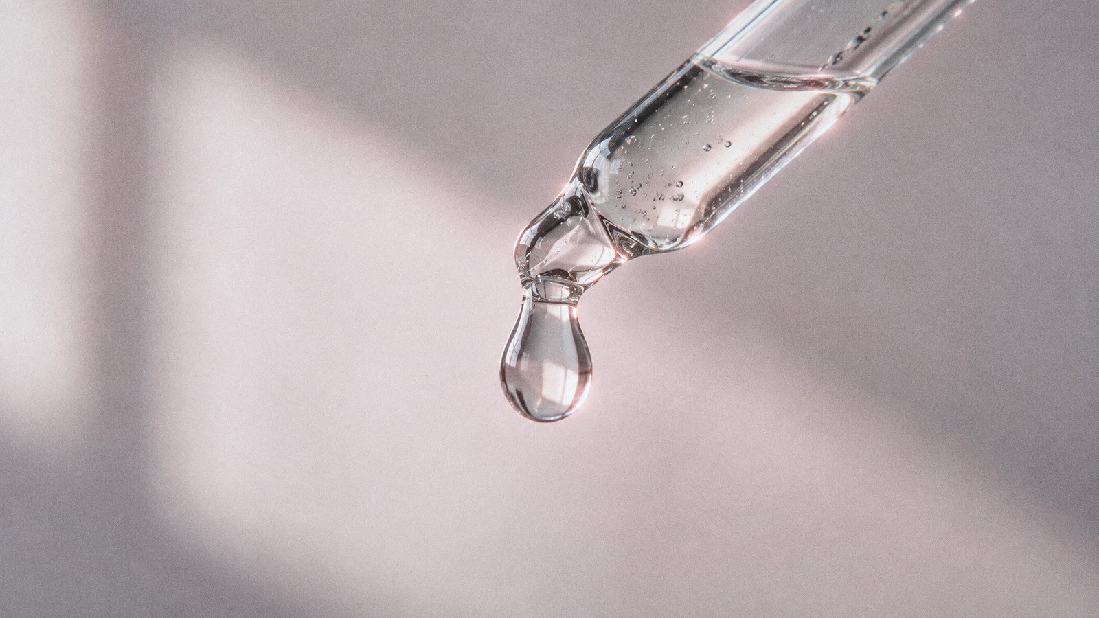 Single drop of clear serum falling from a glass dropper against a soft pink background