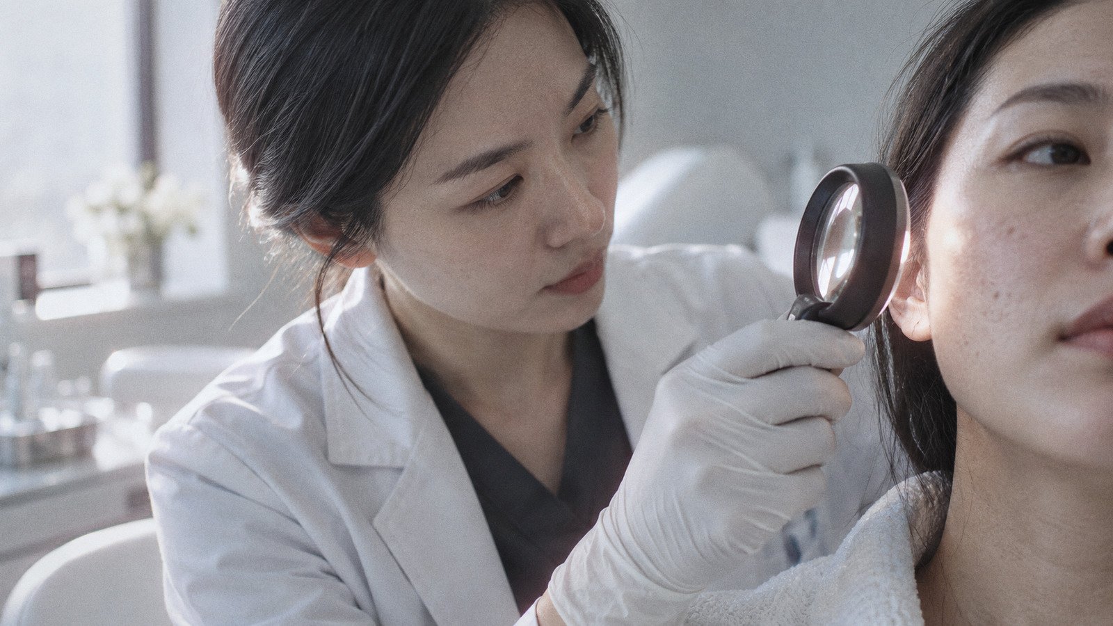 Doctor's gloved hand holding a magnifying lens near a patient's cheek during a skin assessment