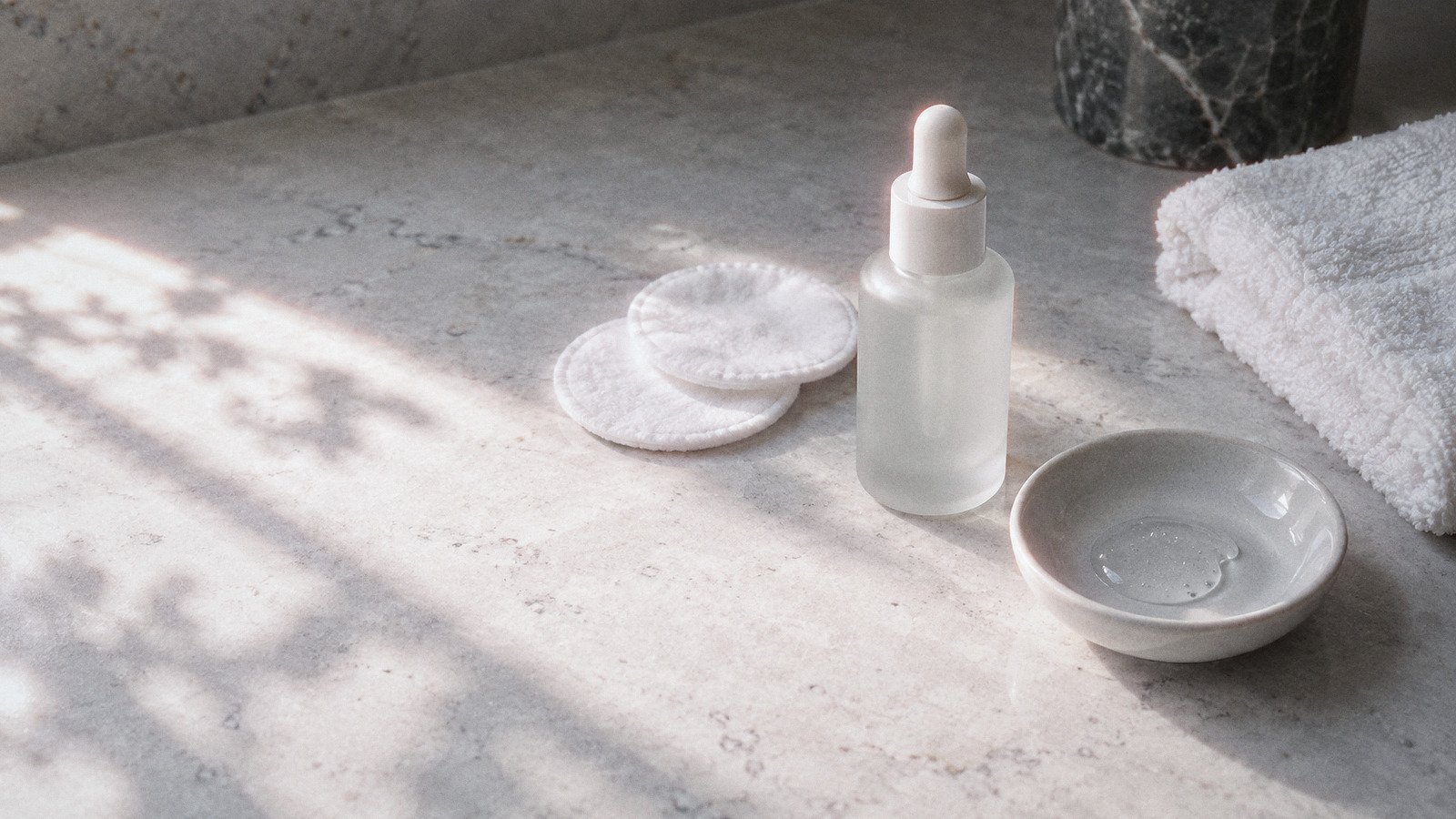 Unlabelled serum bottle, cotton pads, a dish of clear gel and a folded towel on a stone bathroom counter